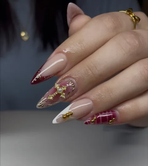 Long stiletto acrylic nails in burgundy, white, and gold with sculpted cross and pearl details by Leah